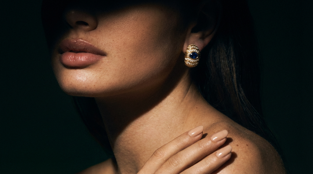 Close-up of a woman wearing a gold earring with a blue gemstone against a dark background