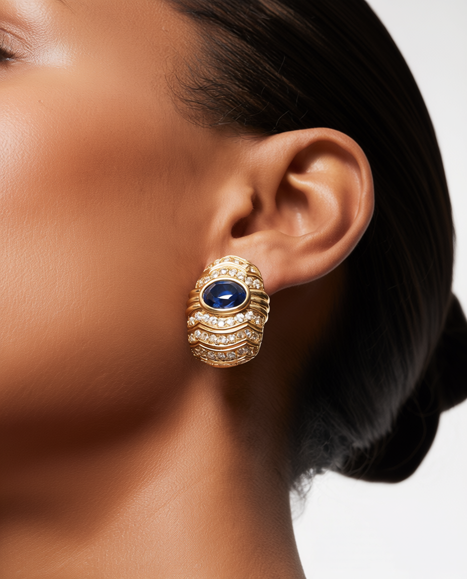 Close-up of a person wearing a gold earring with a blue gemstone on a neutral background