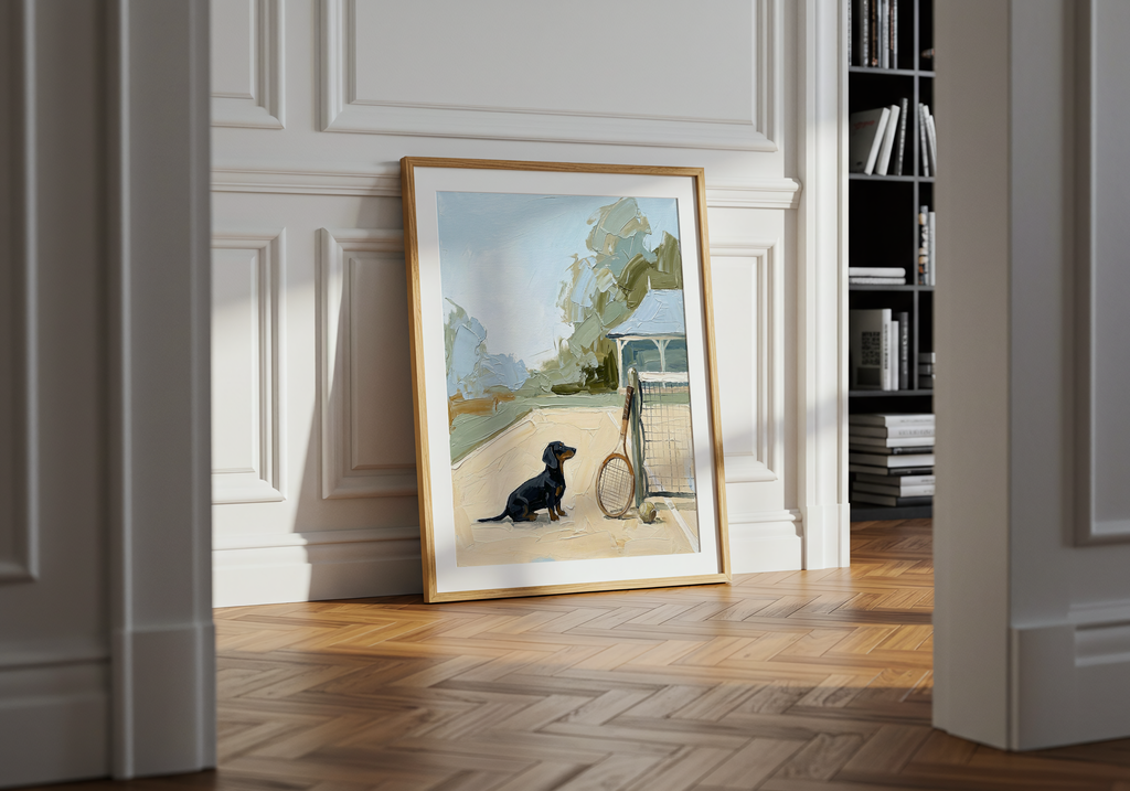 Framed artwork of a dog with a bicycle on a wall in a room with wooden flooring and a bookshelf.
